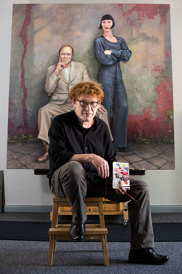 Manfred W. Jürgens sitzt vor einem seiner Gemälde. Er hält zwei Pinsel und eine Mischpalette in der Hand.