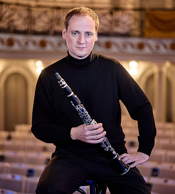 Ein Mann sitzt auf einem Hocker in einem leeren Konzertsaal. Hinter ihm sind viele Sitzreihen. Der Mann hat eine Klarinette in der Hand.