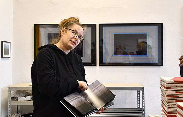Ramona Seyfarth vor ihren Arbeiten. In der Hand hält sie ein aufgeschlagenes Buch. 