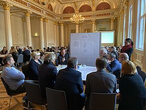 Im Raum stehen zusammengestellte Tische. Darum verteilen sich Frauen und Männer. Vor einem Tisch steht eine Pinnwand zum Rollen.