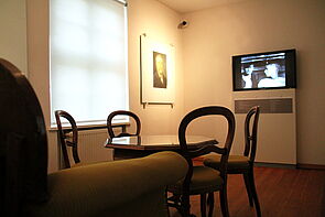 Eine Stube mit Couch, Tisch und Stühlen. An der Wand hängt ein Fernseher, auf dem ein Film läuft. 