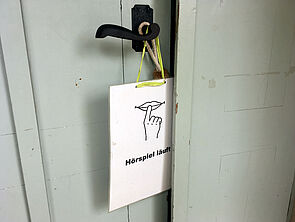 An der Klinke der Eingangstür hängt ein Schild mit einer Zeichnung von einem Finger auf geschlossenen Lippen und der Aufschrift "Hörspiel läuft".