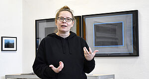 Ramona Seyfarth gestikuliert vor zwei Arbeiten aus ihrer Serie 24 Hours. Fotoarbeiten in Rahmen hängen hinter ihr an der Wand.
