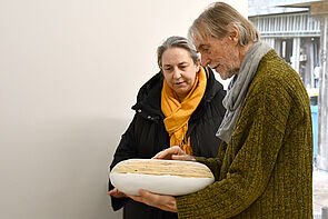 Broder Burow hält eine Holzarbeit im Arm. Hinter ihm eine Frau, die seine Arbeit anschaut.