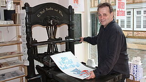 Hans-Hilmar Koch steht vor einer schwarzen Druckmaschine und hält ein bedrucktes Blatt in der Hand.