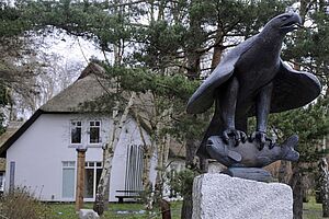 Ein weißes, restgedecktes Haus, umgeben von Wiese und Bäumen. Im Vordergrund steht auf einem weißen Sockel die Büste eines Greifvogels, der einen Fisch in seinen Krallen hält.