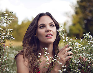 Eine Frau. Sie steht in der Natur. Vor ihr sind kleine, weiße Blumen. 