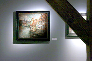 Ein Bild hängt hinter einem Balken. Zu erkennen sind Häuser am Wasser.