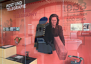In einer Vitrine befinden sich eine Informationswand, ein Posthorn, ein Paket, eine Briefwaage und ein Telefon. 