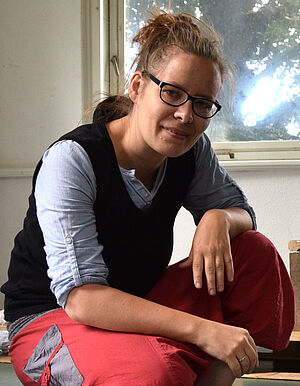 Ramona Seyfarth hockt vor einer weißen Platte. Darauf steht ein transparenter Kinderstuhl mit Lehne. Auf der Sitzfläche steht ein kleiner Spielzeugsoldat. Auf der Platte liegt eine bunte, geschmolzene Masse.