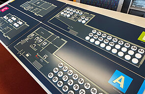 Auf einem breiten, hüfthohen Brett werden die Räume der Bibliothek abgebildet. Es enthält Informationen in Form von Piktogrammen, Blindenschrift und großen, weißen Knöpfen mit Zahlen.