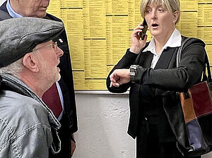 Eine Frau. Sie steht vor einem Fahrplan der Bahn. Sie telefoniert und schaut auf ihre Uhr. Vor ihr stehen zwei Männer.