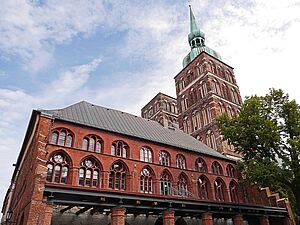 Eine Backsteinfassade. Zwei Fensterreihen mit jeweils neun Fenstern. Davor steht ein Baum. Dahinter ragt der Turm der Stralsunder Nikolai-Kirche empor.