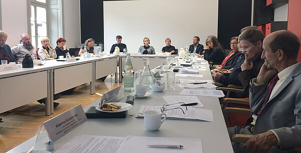 Ein Konferenztisch in U-Form. An ihm sitzen 14 Frauen und Männer. Auf dem Tisch befinden sich bedruckte Blätter, Stifte, Kaffeetassen, Gebäck, Getränkeflaschen und Namensschilder.