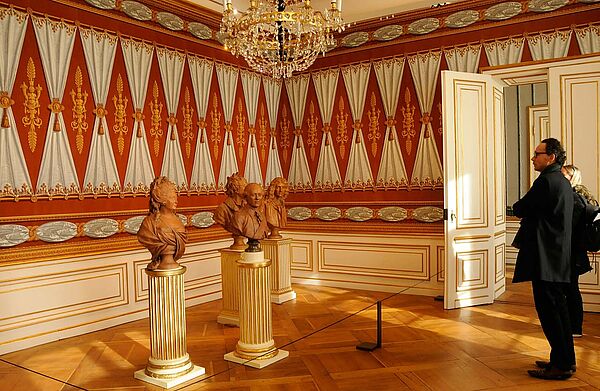 Eine rot-weiß-gold verzierte Wand. Davor stehen vier terrakottafarbene Büsten.