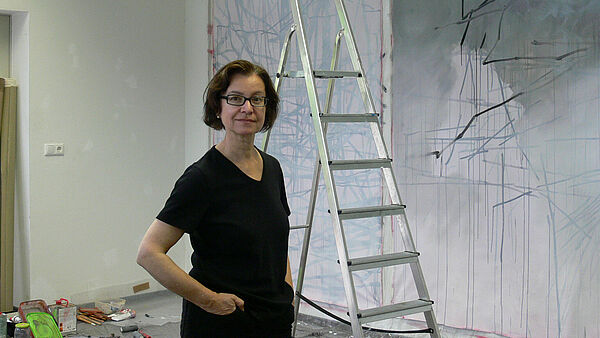 Eine Frau steht in einem Raum. Sie hat die Hände in den Taschen. Hinter ihr steht eine Leiter. An den Wänden hängen raumhohe Zeichnungen.