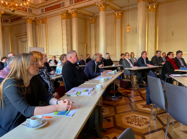 Ein historischer Saal mit Säulen und goldfarbenen Verzierungen an den Wänden. Verteilt auf drei Tischreihen sitzen rund 20 Frauen und Männer.