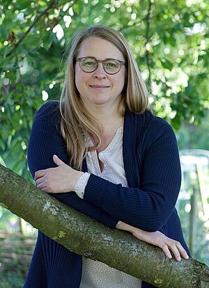 Porträt einer Frau. Sie steht an einem Baum und lehnt auf einem Ast.