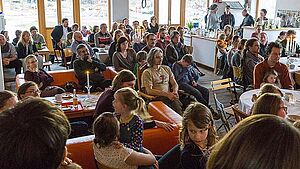 Ein Raum voller Menschen. Sie sitzen an Tischen.