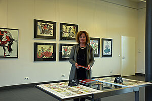 Eine Frau steht in einem Raum. Sie hält ein Heft in den Händen. Hinter ihr, an der Wand, hängen Bilder.