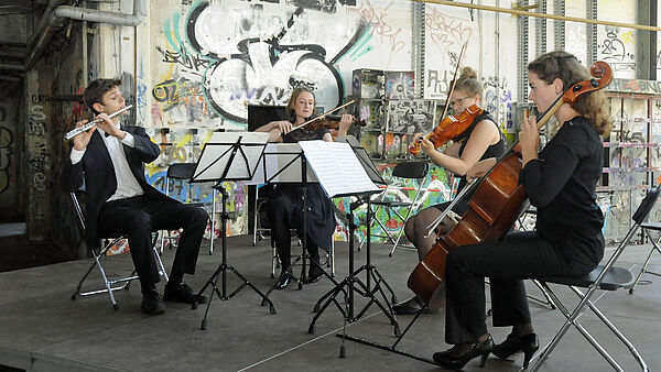 Ein musikalisches Quartett sitzt vor einer Graffiti besprühten Wand und spielt Querflöte, Geige und Cello.
