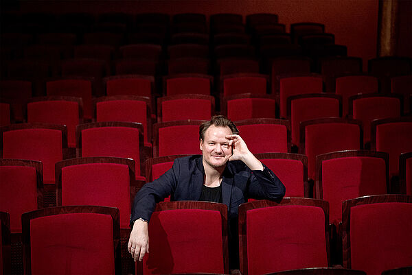 Daniel Geiss sitzt inmitten der roten Stuhlreihen. Eine Hand hängt über der Lehne, auf die andere stützt er seinen Kopf.