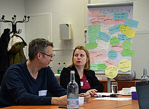 Ein Mann und eine Frau sitzen in einem der Workshops an einem Tisch und unterhalten sich. Hinter ihnen ein Aufsteller mit farbigen Zetteln. Darauf stehen Gedanken und Ideen zum Thema "Kulturland MV".