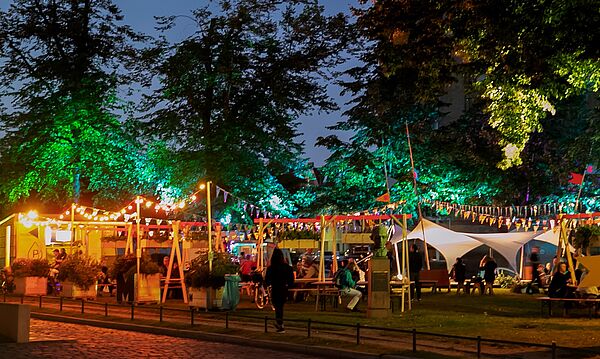 Menschen sitzen auf Bänken. Der Platz ist bunt beleuchtet.