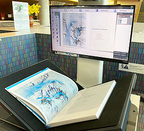 Das Buch „Geografie der Antarktis“ liegt geöffnet auf dem Scanner. Die zu scannende Seite wird auf dem dazugehörigen Monitor angezeigt.