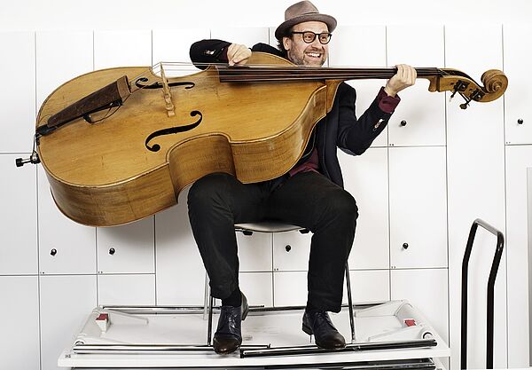 Ein Mann. Der Mann sitzt in einem weißen Raum. Er sitzt auf einem Stuhl und hält einen Kontrabass. 