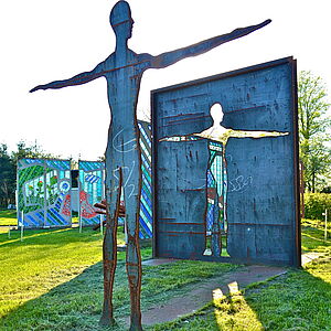 Befreiung von Max Schmelcher Eine Skulptur in Form eines Menschen. Sie steht mit ausgebreiteten Armen auf einer Wiese. Dahinter steht ein Rechteck, aus dem sie ausgeschnitten zu sein scheint.