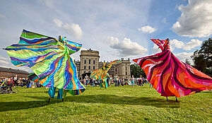 Drei Menschen auf Stelzen. Sie tragen bunte Kleider und tanzen auf einer Wiese. Im Hintergrund befinden sich Viele Menschen und das Schloss von Ludwigslust.