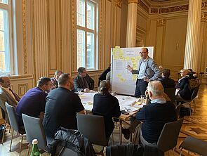 Ein Tisch voller Zettel. Darum verteilen sich neun Frauen und Männer. Ein Mann steht am Tisch neben einer Magnetwand, an der ebenfalls viele Zettel hängen.